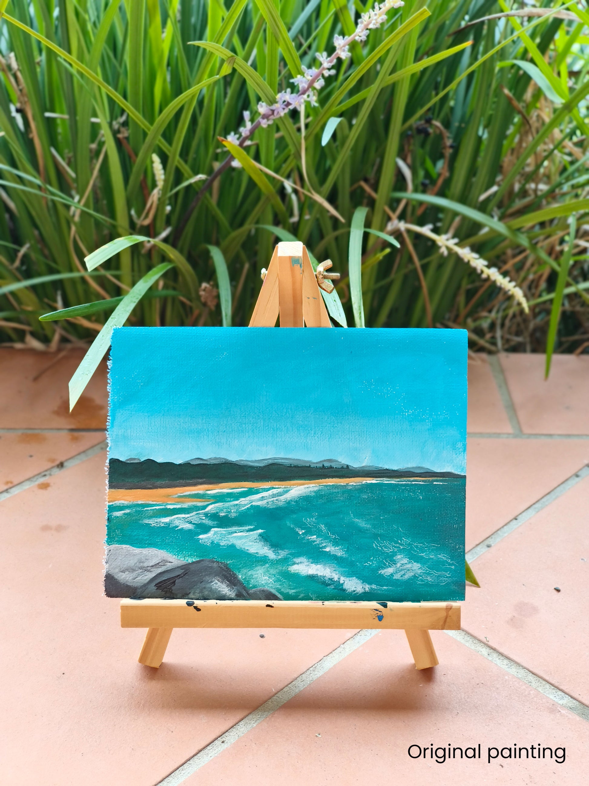 Small painting of a beach scene on an easel with greenery in the background. the caption in the photo says "Original painting", showing that this is the original painting the prints were created from.