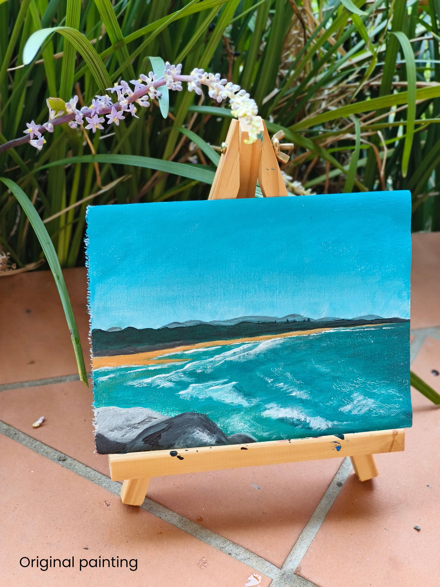 Small painting of a beach scene on an easel with greenery in the background. the caption in the photo says "Original painting", showing that this is the original painting the prints were created from.