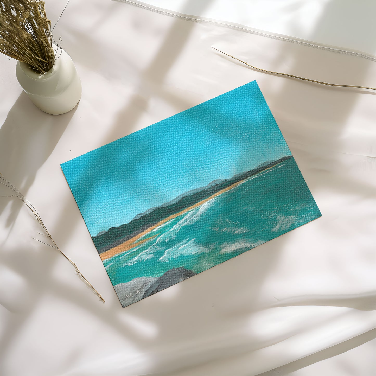Painting of a coastal scene at North Wall Beach, Coffs Harbour, with blue water and waves and clear sky. Set upon a white table near a vase of dried flowers, natural sunlight and shadows pouring in.