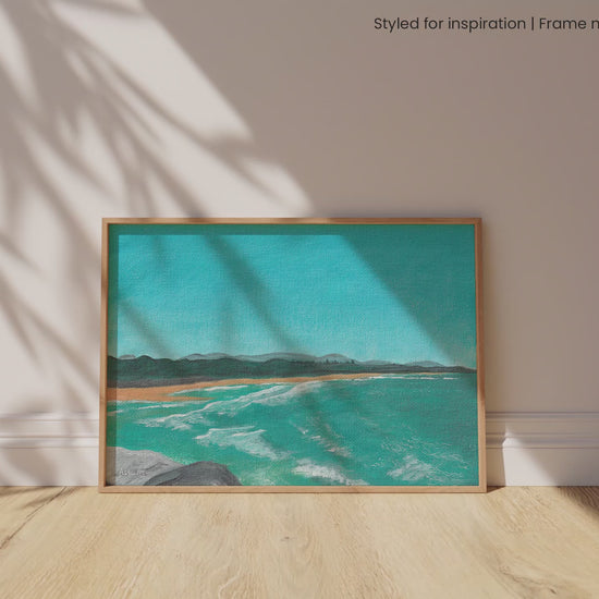 Framed painting of a coastal scene at North Wall Beach, Coffs Harbour, with blue water and waves and clear sky. It is leaning against a white wall, set upon a timber floor with natural light pouring in, and shadows dancing across the scene.