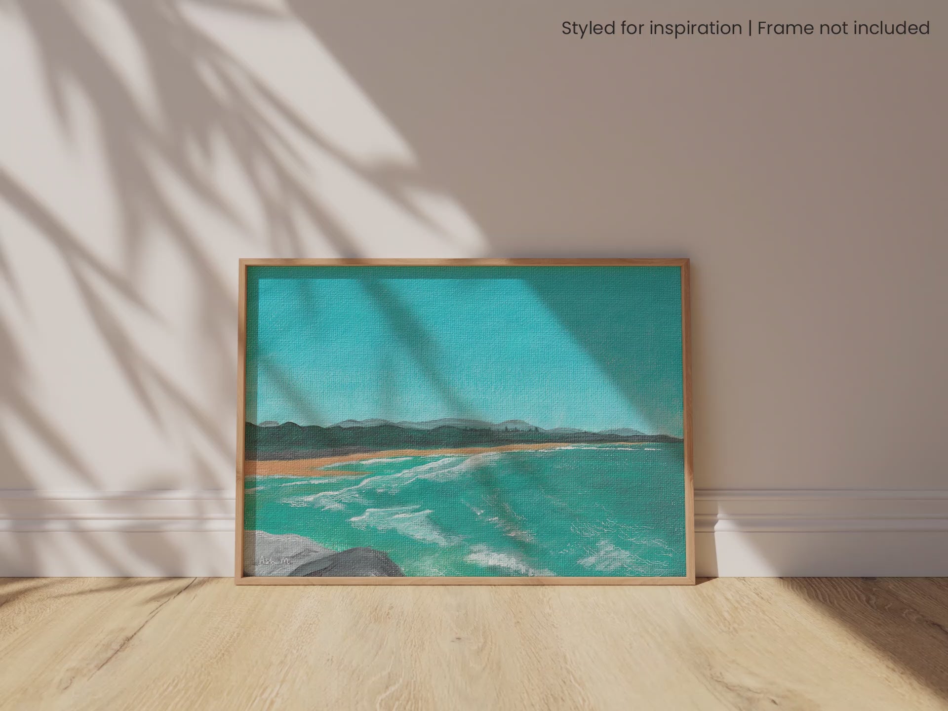 Framed painting of a coastal scene at North Wall Beach, Coffs Harbour, with blue water and waves and clear sky. It is leaning against a white wall, set upon a timber floor with natural light pouring in, and shadows dancing across the scene.